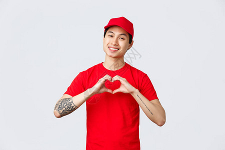 穿着红帽和T恤公司制服的英俊亚洲信使友好地展示心脏手势图片