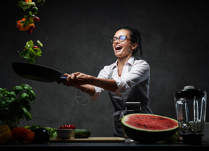 情感成熟的女厨师将切碎的蔬菜从锅里扔出健康食品概念黑暗背景背景图片