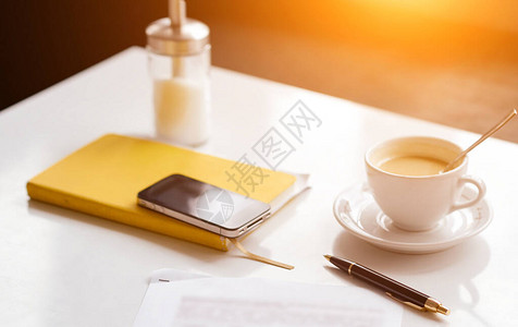 白色桌子上的黄色笔记电话杯咖啡笔和纸片图片