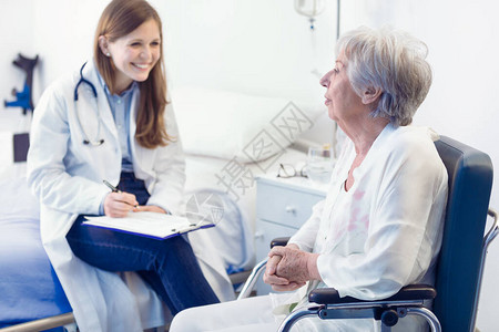 护士或医生在病房里为坐在轮椅上的微笑老妇人写病历图片