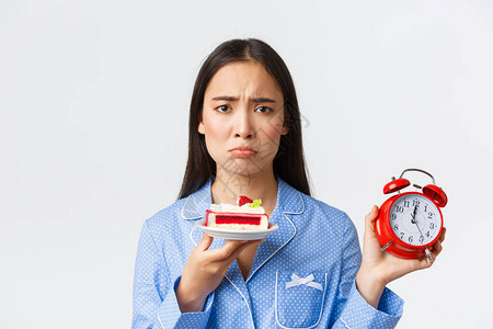 穿着睡衣的心烦意乱的可爱噘嘴亚洲女孩的特写镜头显示时钟图片