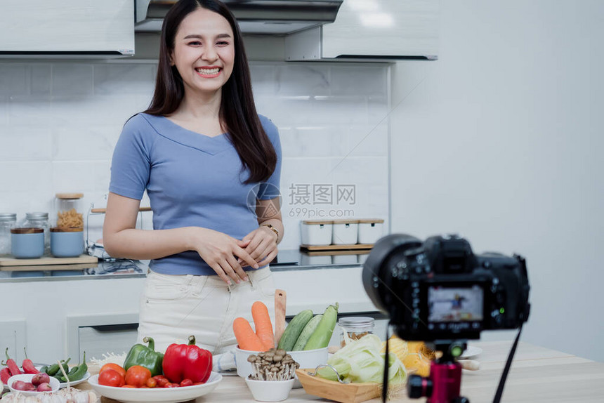 社交媒体概念一个快乐的女人站在厨房里用相机在线录制视频快乐的亚洲女人vlogger直播在线视频教学图片