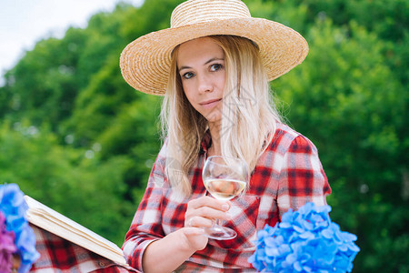 穿着红色格子连衣裙和帽子的女孩坐在白色针织野餐毯上看书和喝酒图片
