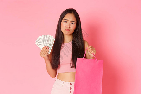 失望闷不乐的亚洲女孩带着钱和购物袋看起来不开心的肖像图片