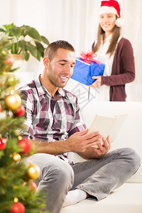 年轻漂亮的男人在看书而他的女朋友却拿着圣诞礼物她想给他一个惊图片