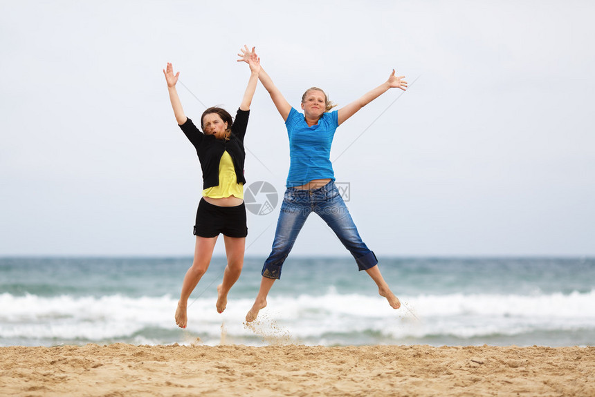 两个快乐的女孩在海滩上与海和无云的天空对着跳浅的田地深处图片