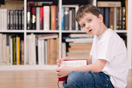 图书馆里有红书的孩子看着相机他盘腿坐在地板上身着白色T恤图片