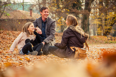 快乐的三口之家在秋日下跪在树叶覆盖的公园地面上图片