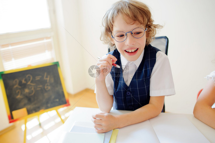 快乐的小学生戴着眼镜坐在课桌旁是一所小学的弟子在男孩圆珠笔的手中课桌上放着抄写本靠近课桌图片