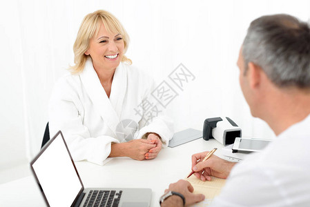 我觉得自己很棒快乐的高级女人正在与男医生交谈并微笑她坐在白色浴袍男图片