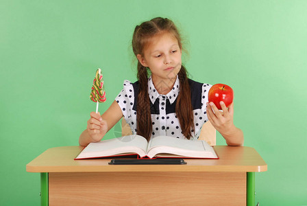 穿着校服的女孩坐在书桌上选择糖果或苹果在图片