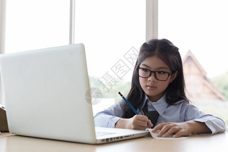 快乐的亚洲年轻女孩坐在桌边学习在线学习在家用笔记本电脑写笔记背景图片