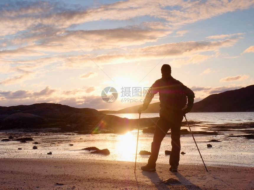 沃克在海上观看阳光明媚的春天黎明背着包的徒步旅行者站在沙滩上太阳光线反射在水和湿沙中徒步旅行者享受令人惊叹的日图片
