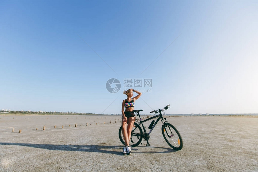 身着五彩西服的金发强壮女人站在沙漠地区的一辆自行车旁图片