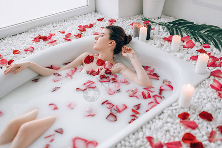 花瓣焕肤年轻女子躺在浴缸与泡沫玫瑰花瓣和燃烧蜡烛装饰背景