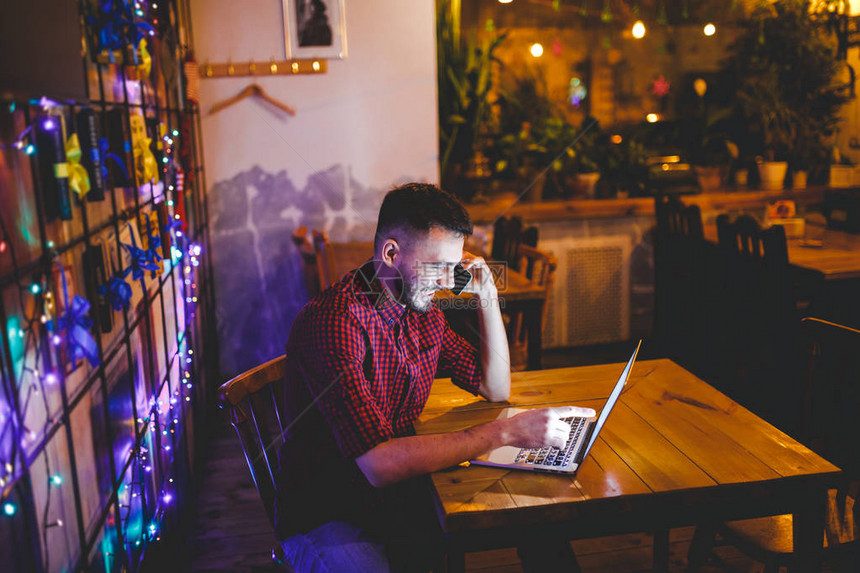 年轻英俊的白种人留着胡子戴着衬衫带着的微笑在笔记本电脑后面工作坐在木桌旁的键盘上在手机上使用电话晚上在图片