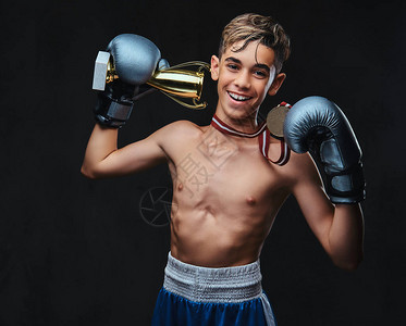 快乐的年轻的无上衣的拳击手戴着手套拿着冠军杯和金牌在黑暗下被隔离背景图片