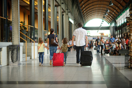 年轻的父母带着两个孩子在车站一家人穿过一个大等候室都牵手爸妈带着大手提箱背着包的男背景图片