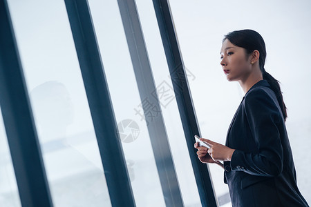 拿着手机的商务女士站在玻璃窗前图片