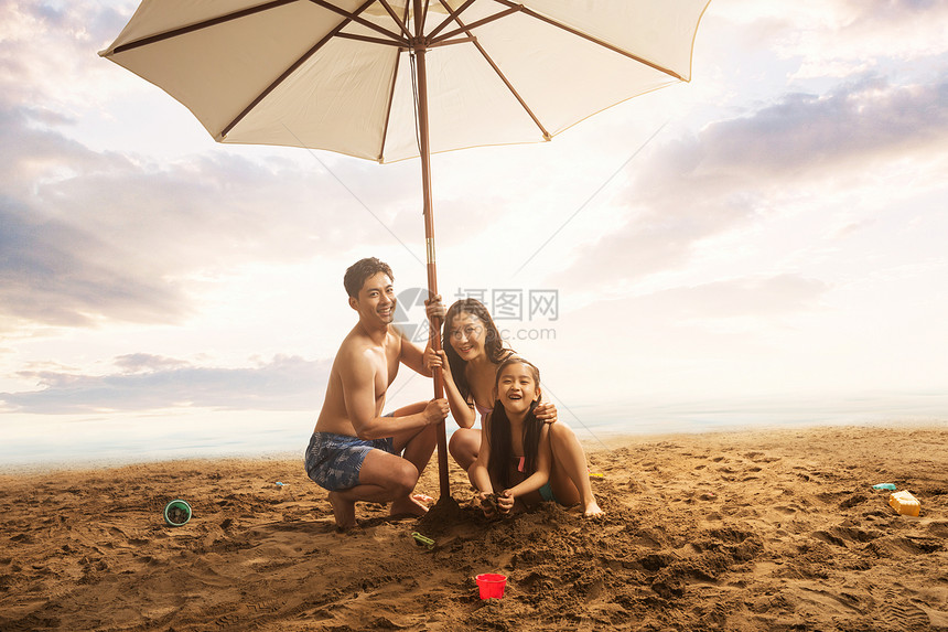 海滩上躲在遮阳伞下的三口之家图片