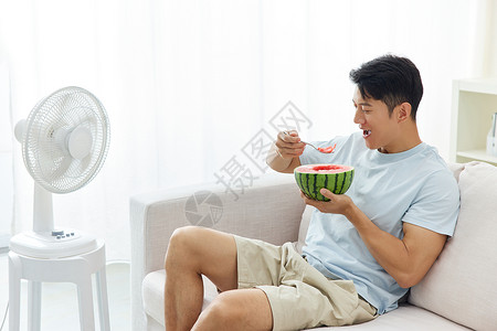 男人靠在沙发上吃西瓜吹风扇降温高清图片素材