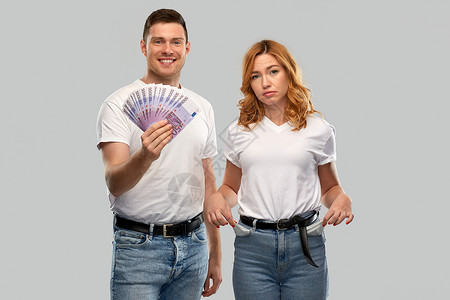 金融,储蓄夫妇快乐的年轻人穿着白色T恤,着欧元的钱悲伤的女人,口袋空空,背景灰色夫妇有欧元空口袋图片