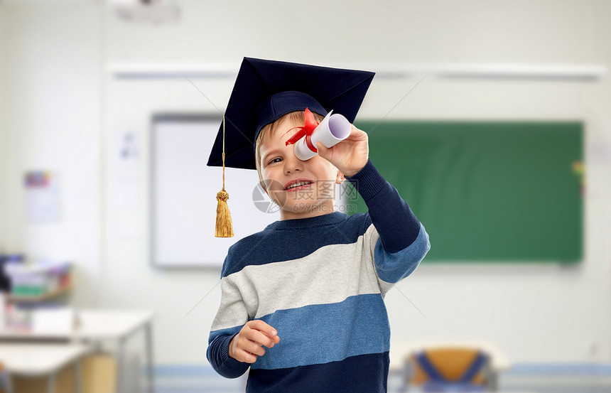学校,教育学快乐的小男孩戴着学士帽或黑板,透过文凭的课堂背景灰泥板上的小男孩看着文凭图片