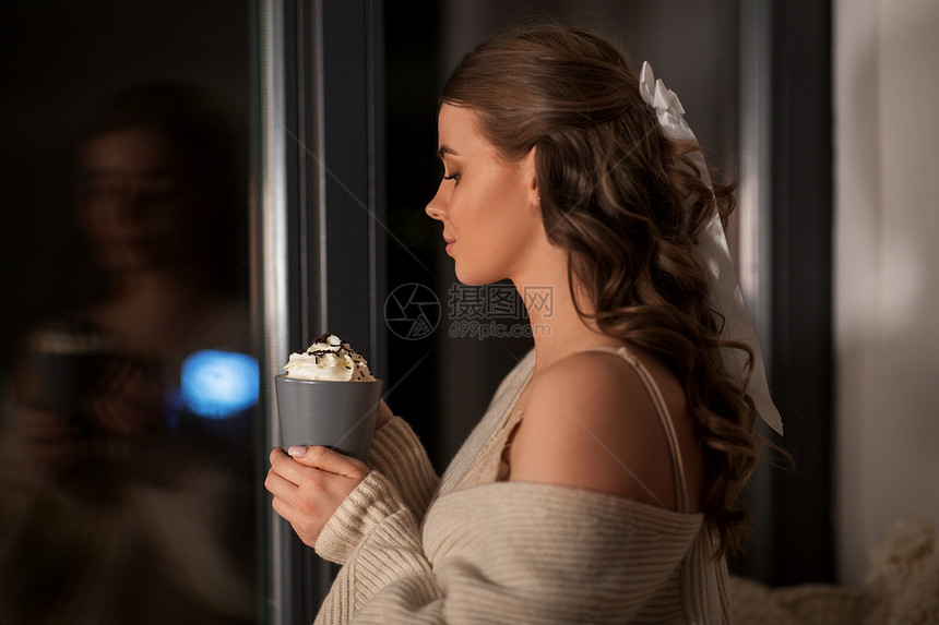 冬天,舒适人的年轻的女人穿着套衫,晚上窗户上着奶油晚上着奶油的杯子的女人图片