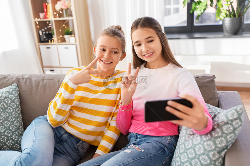 人,技术友谊的快乐的十几岁女孩自拍与智能手机坐沙发上家快乐的女孩家里用智能手机自拍图片