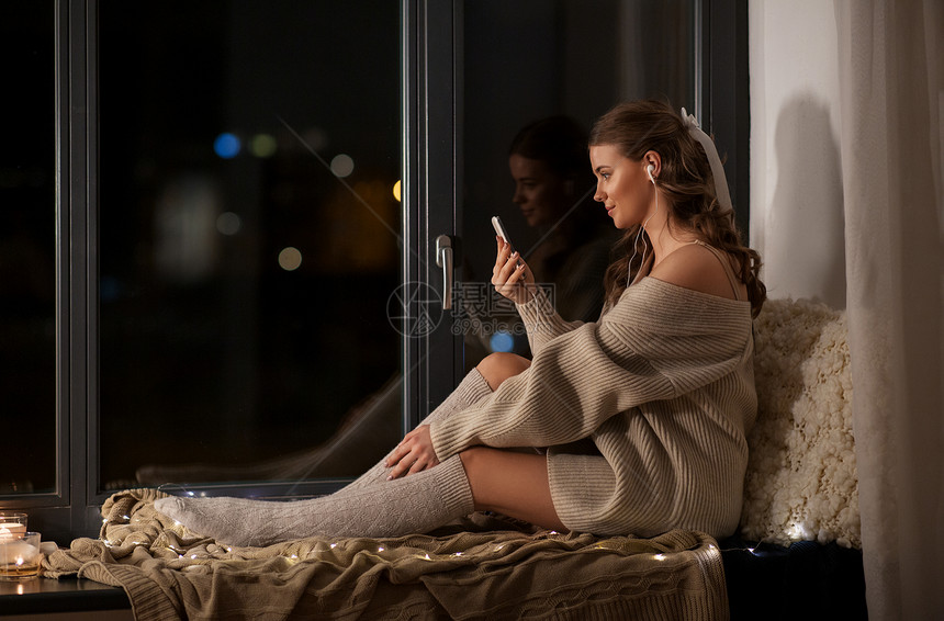技术,舒适人的年轻的女人穿着套衫,带着智能手机耳机,听着音乐坐家里的窗台上家里有智能手机耳机的女人图片