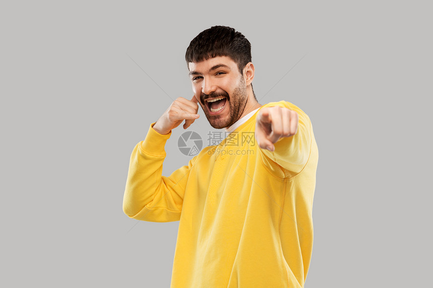 情感人的快乐的微笑年轻人穿着黄色运动衫,打着电话手势,手指指向灰色背景的相机男人打电话手势指指点点图片