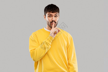 秘密人们的微笑的年轻人穿着黄色运动衫,灰色背景上用手指嘴唇上做着沉默的手势年轻人用手指嘴唇上做着沉默的手势图片