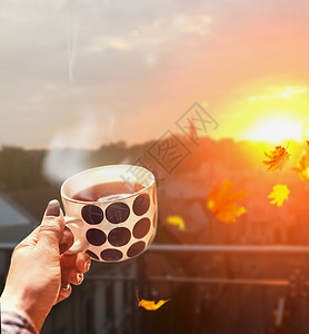 女人手着杯子,秋天的城市背景下,用热饮料蒸汽,带着飞扬的树叶夕阳的光芒图片