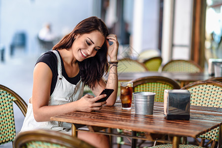随便什么微笑的年轻女人,蓝色的眼睛坐城市咖啡馆用智能手机棕色波浪发型的女孩穿白色牛仔连衣裙生活方式的背景