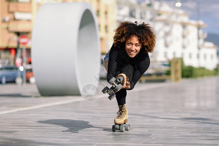 留音机年轻合身的黑人妇女溜冰鞋骑户外的城市街道上阳光明媚的日子里,微笑的女孩留着AFRO的发型骑着溜冰鞋的黑人女人城市街背景