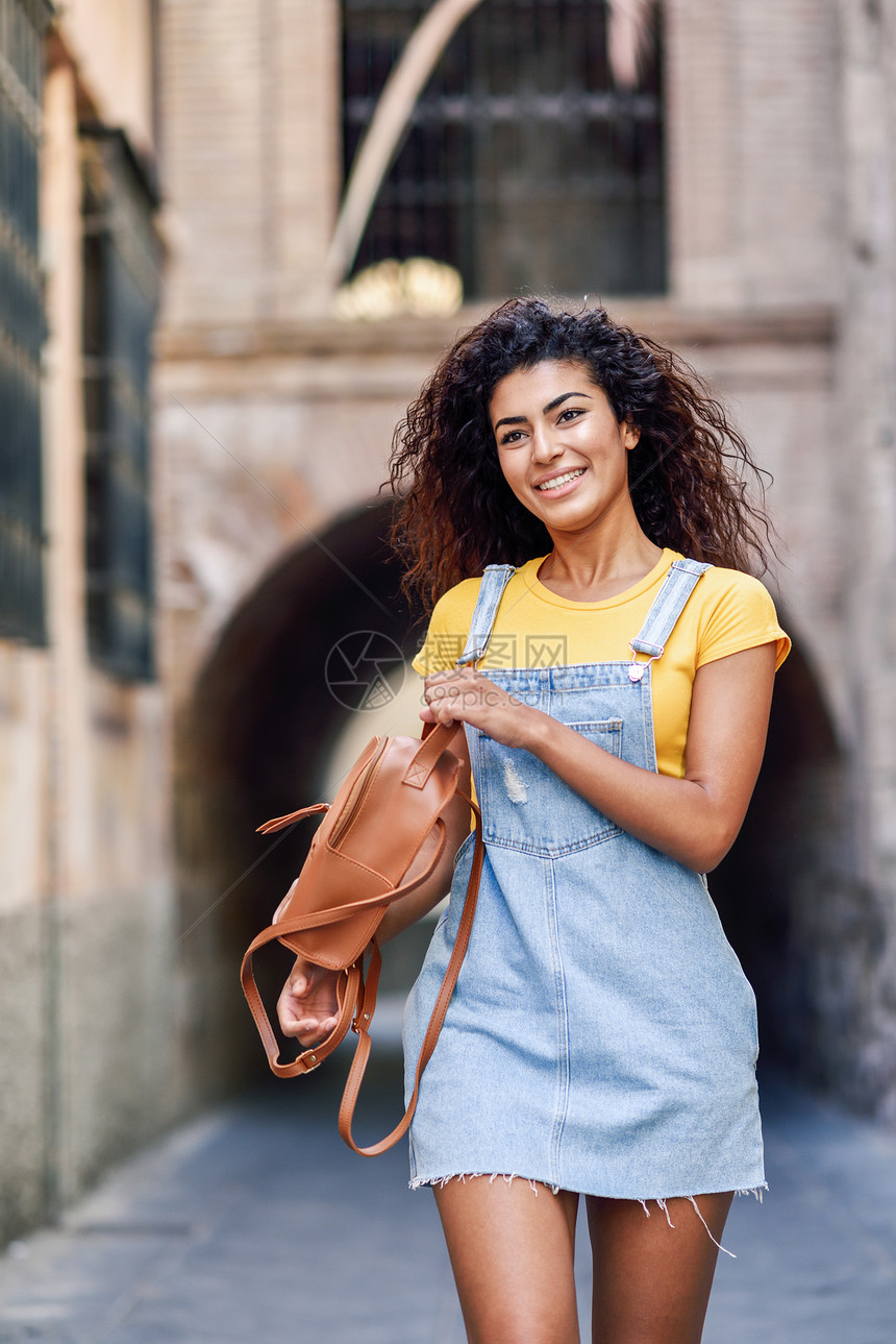 年轻的阿拉伯旅游女,留着卷曲的发型街上穿便装的阿拉伯旅行者女孩快乐的女穿着黄色T恤城市背景的牛仔连衣裙年轻的黑图片