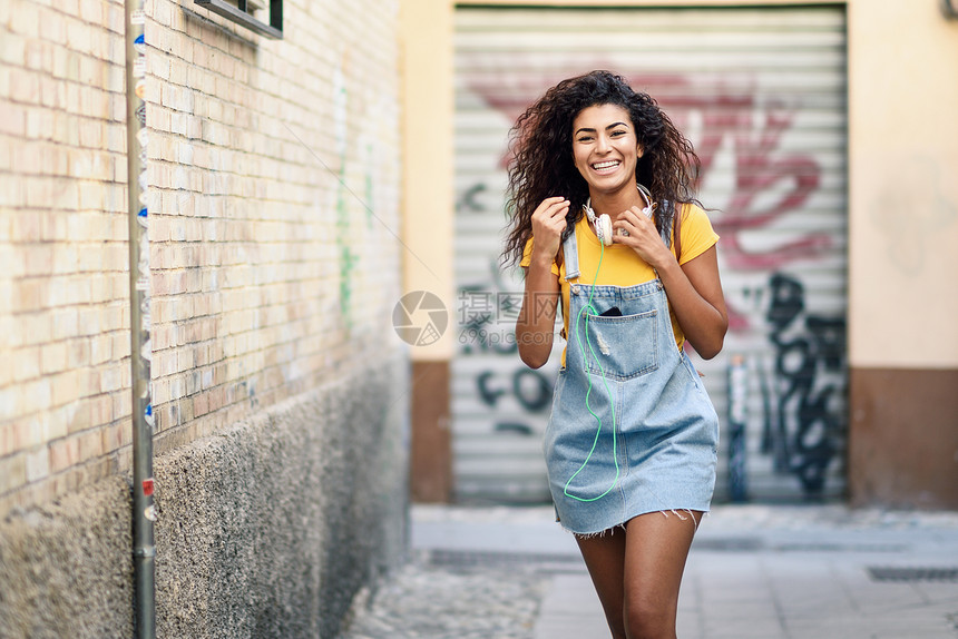 年轻的阿拉伯女人带着耳机黑色卷发走户外快乐的女孩穿着黄色的T恤城市背景的牛仔连衣裙带着耳机走户外的年轻非洲女人图片