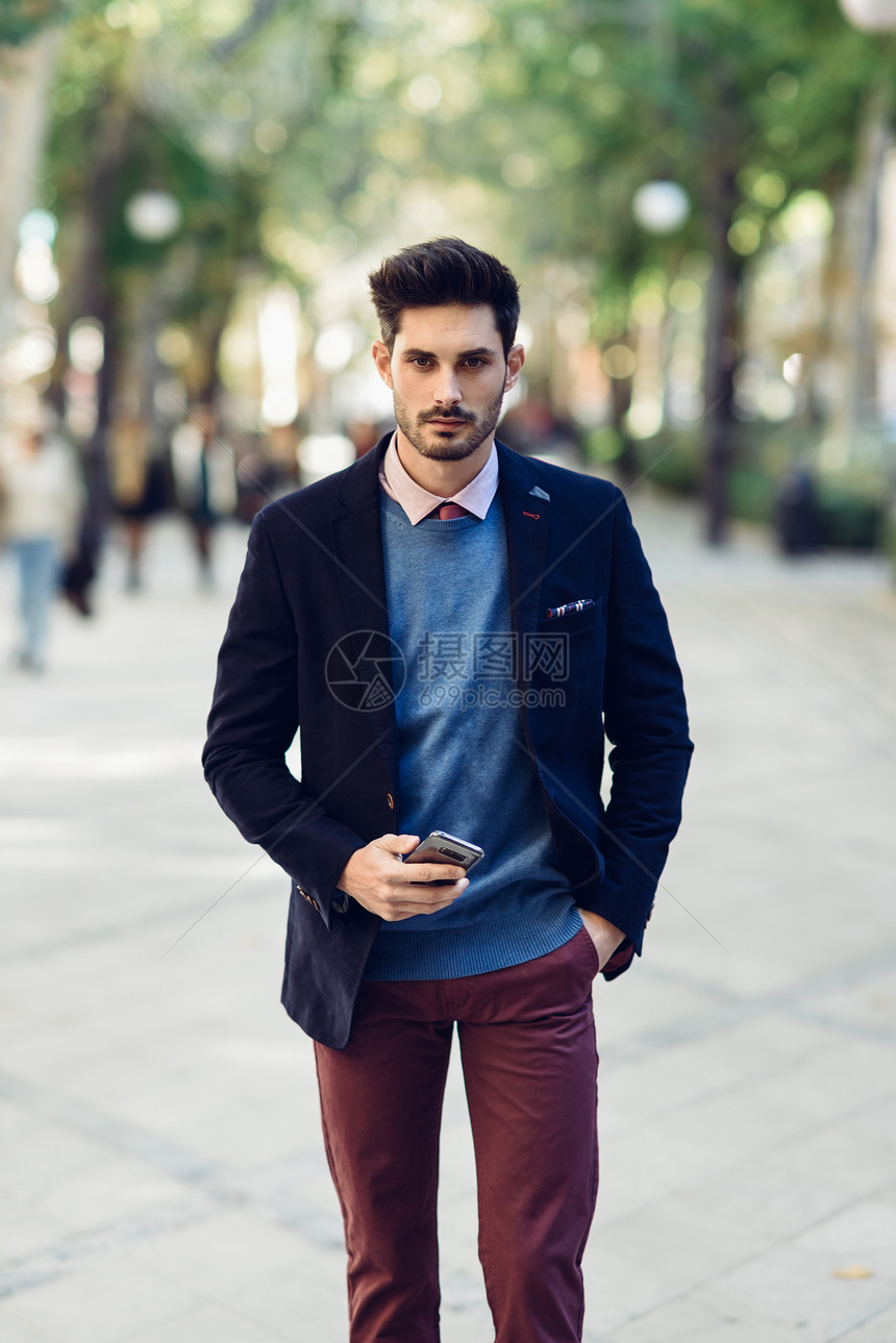 街上有魅力的男人穿着英国优雅的西装,手里着智能手机年轻的留胡子的商人,城市背景下有现代发型街上的男人穿着正装,手里着图片