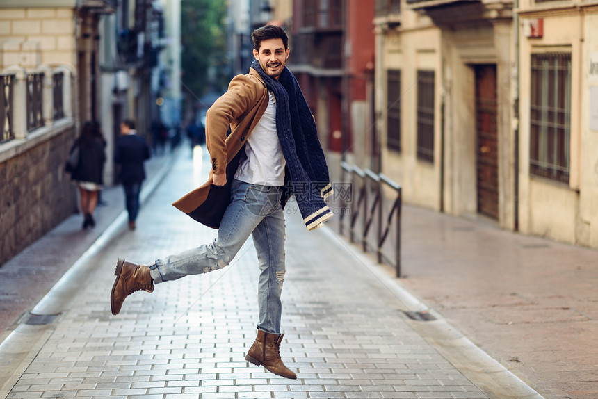年轻的快乐男人穿着冬天的衣服街上跳跃年轻的留胡子的家伙,现代发型,外套,围巾,蓝色牛仔裤t恤城市背景年轻的快乐的男人图片