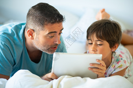 中年父亲她八岁的女儿卧室里用数字平板电脑有趣的爸爸可爱的小女孩床上玩得很开心中年父亲她八岁的女儿卧室里用数背景