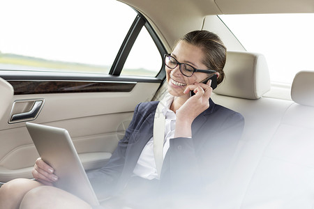 微笑的女商人智能手机上交谈,同时汽车上用数字平板电脑图片