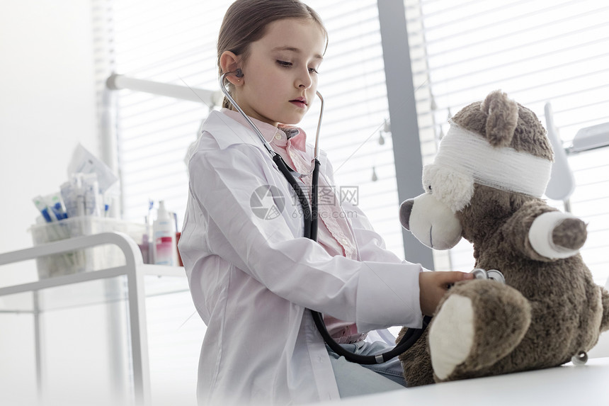 医院用听诊器检查泰迪熊的时候,穿着实验室大衣模仿医生的女孩图片