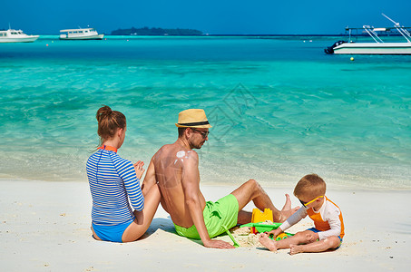 保护海洋孩子海滩上的家人,三岁男孩的轻夫妇女男背部涂防晒霜马尔代夫的暑假背景