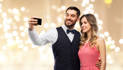 技术人们的快乐的夫妇穿着派服装,用智能手机米色背景下用节日灯自拍幸福的夫妇用智能手机自拍背景图片