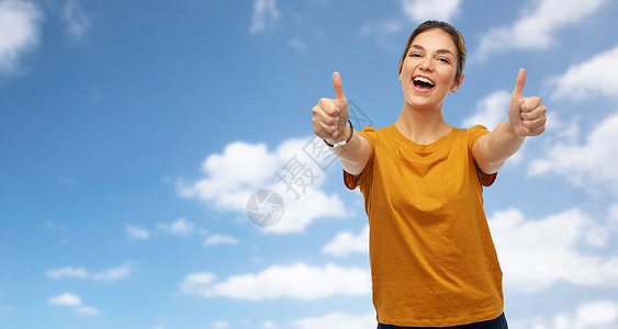 蓝色女孩手势人的快乐微笑的轻女人十几岁的女孩穿着橙色的t恤,蓝天云彩的背景上竖大拇指穿着T恤的女人十几岁的女孩竖背景