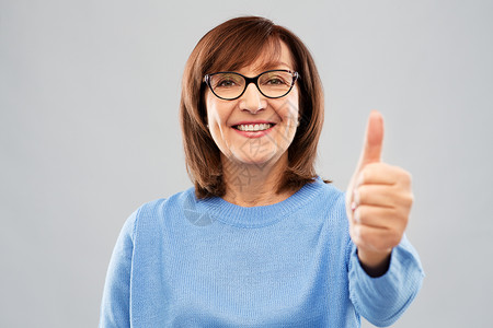 手势,视觉老人的微笑的老妇女眼镜上的肖像,灰色背景上竖大拇指老妇女的肖像竖大拇指背景图片