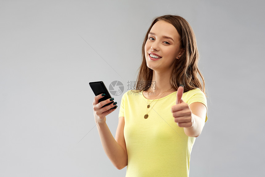 技术人的微笑的轻妇女十几岁的女孩穿着空白的黄色T恤,智能手机拇指灰色背景上智能手机的十几岁女孩竖大拇指图片