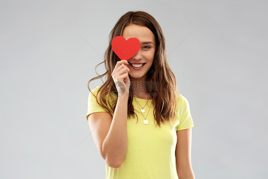 情人节人们的微笑的轻女人十几岁的女孩穿着T恤,用红色的心遮住她的眼睛,灰色的背景微笑的十几岁女孩用红心遮住图片