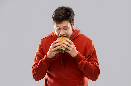 快餐人们的饥饿的轻人穿着红色连帽衫吃汉堡包灰色背景饥饿的轻人吃汉堡包背景图片