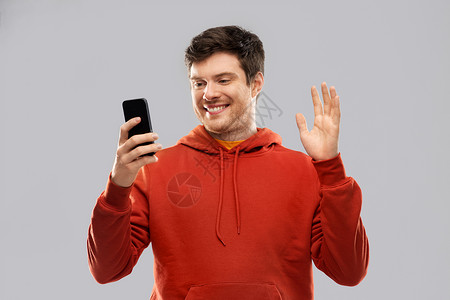 红色连帽衫技术,沟通人的微笑的轻人穿着红色帽衫,智能手机自拍灰色背景上进行视频通话男人用智能手机自拍视频通话背景